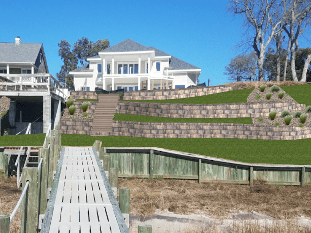A modern white house with tiered landscaping and a wooden walkway leading to the waterfront, surrounded by trees and clear blue sky.