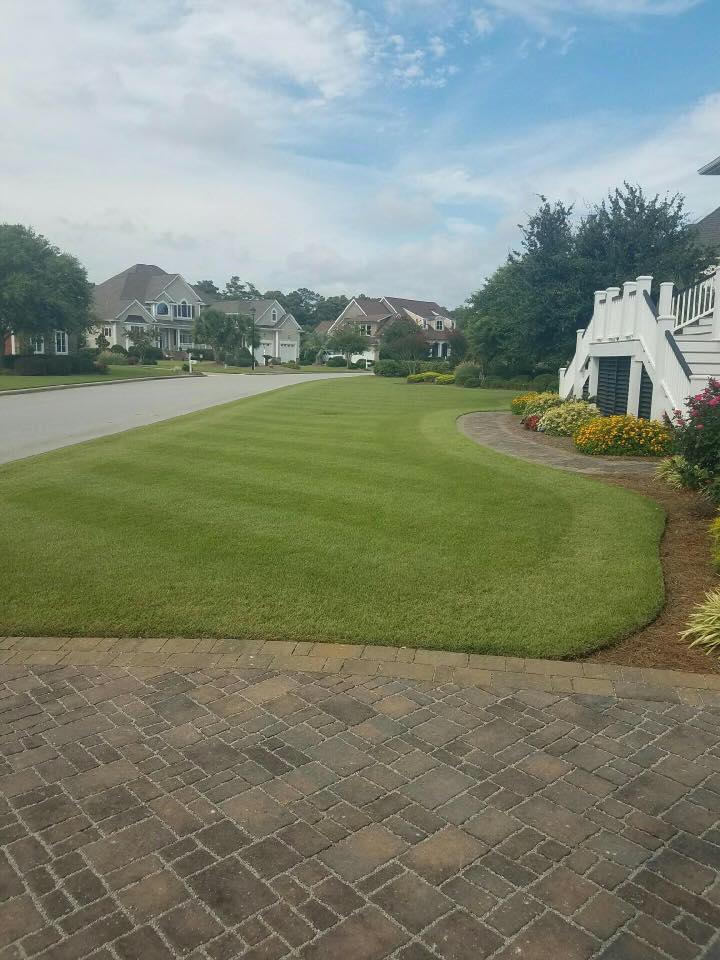 A suburban street with manicured lawns, brick walkway, and two-story houses under a partly cloudy sky. No people or notable landmarks visible.