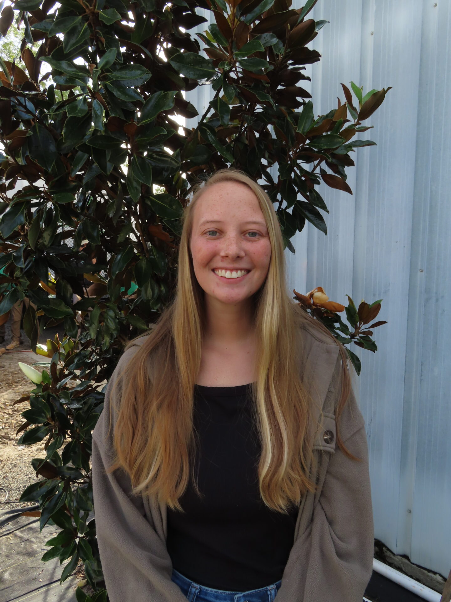 A person with long hair smiles in front of a leafy plant outdoors against a metal wall.