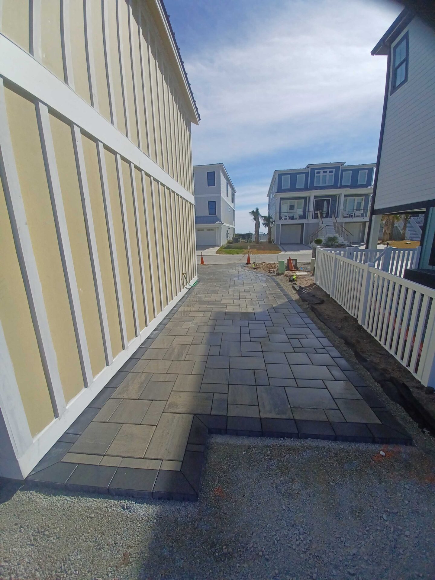 A newly paved pathway between modern houses leads to more dwellings in a coastal residential neighborhood. Bright sky overhead.
