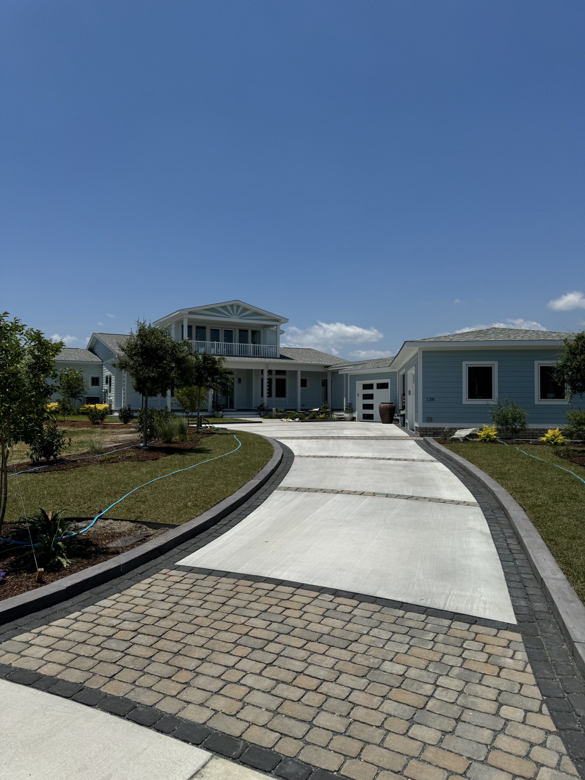 A modern, light blue house with a long driveway, surrounded by landscaped gardens and clear skies, creating an inviting suburban scene.