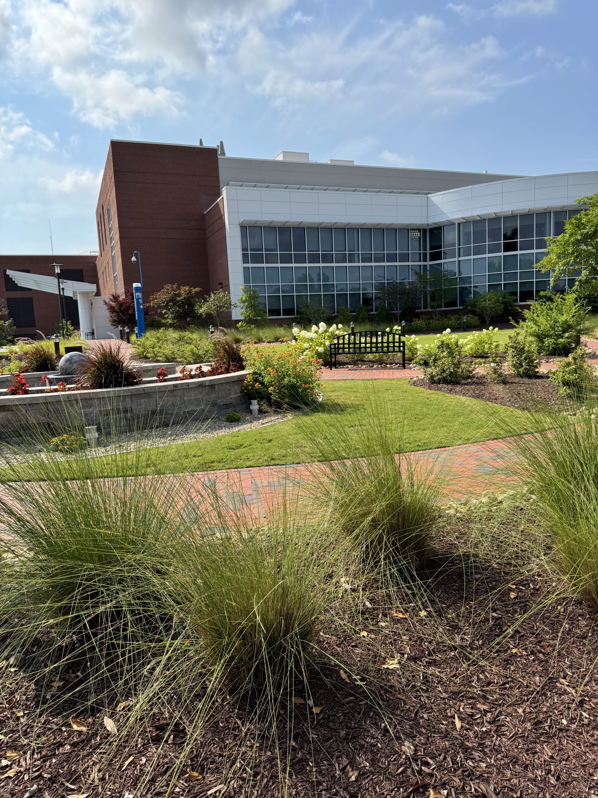 A modern building with red brick and white facade, surrounded by landscaped gardens and pathways under a clear blue sky.