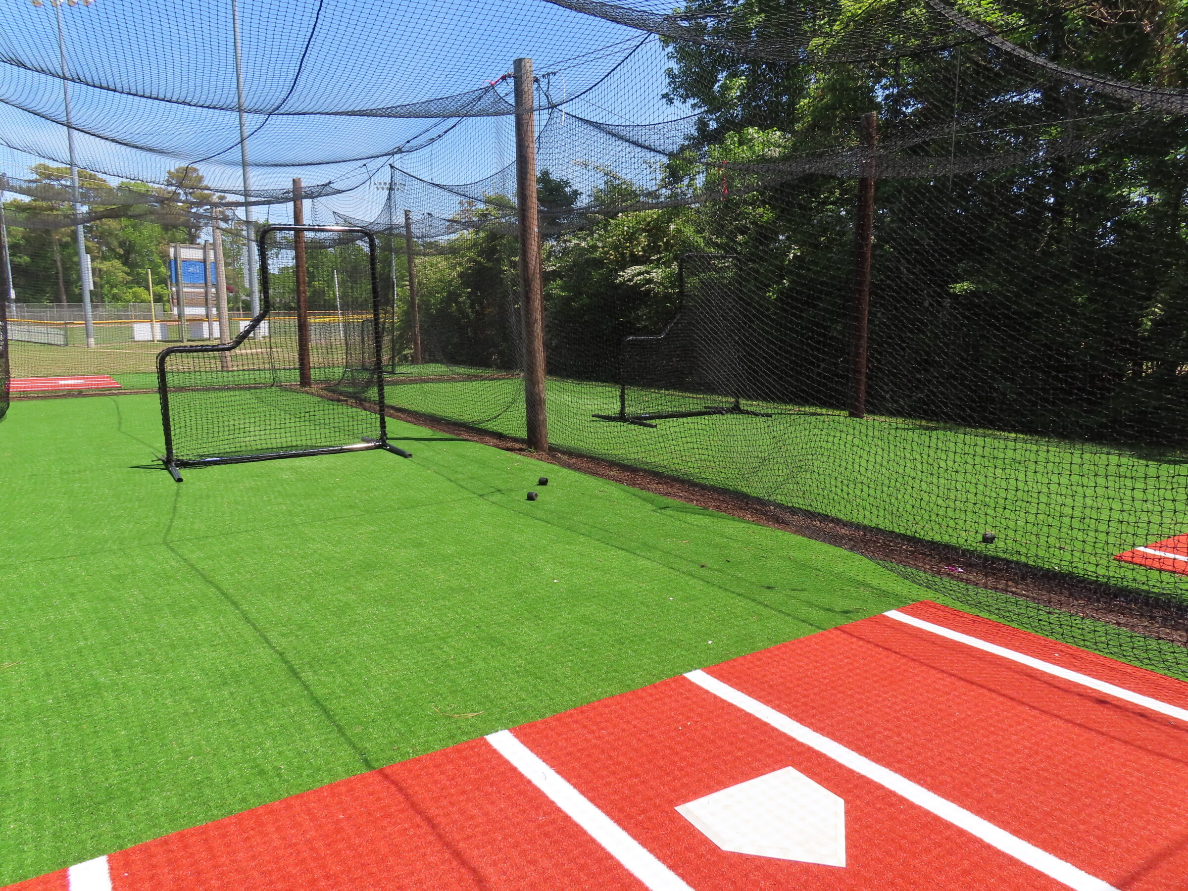 A baseball batting cage with astroturf and nets under a clear sky, surrounded by trees and a fence in the background.