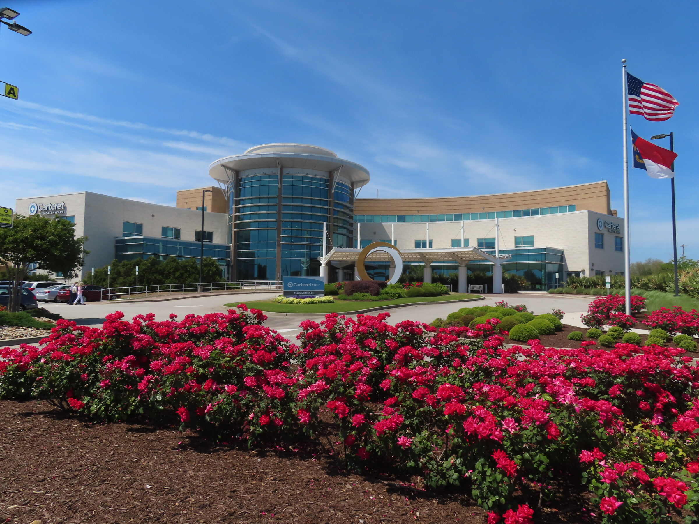 Modern healthcare building with rounded entrance, surrounded by vibrant roses and flags. Two people walk towards the main entrance under a clear sky.