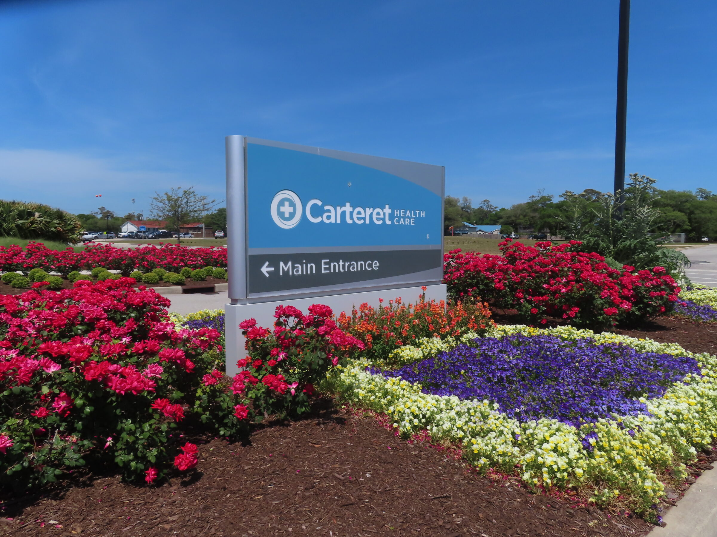 A healthcare entrance sign surrounded by vibrant red, purple, and yellow flowers under a clear blue sky, with trees in the background.