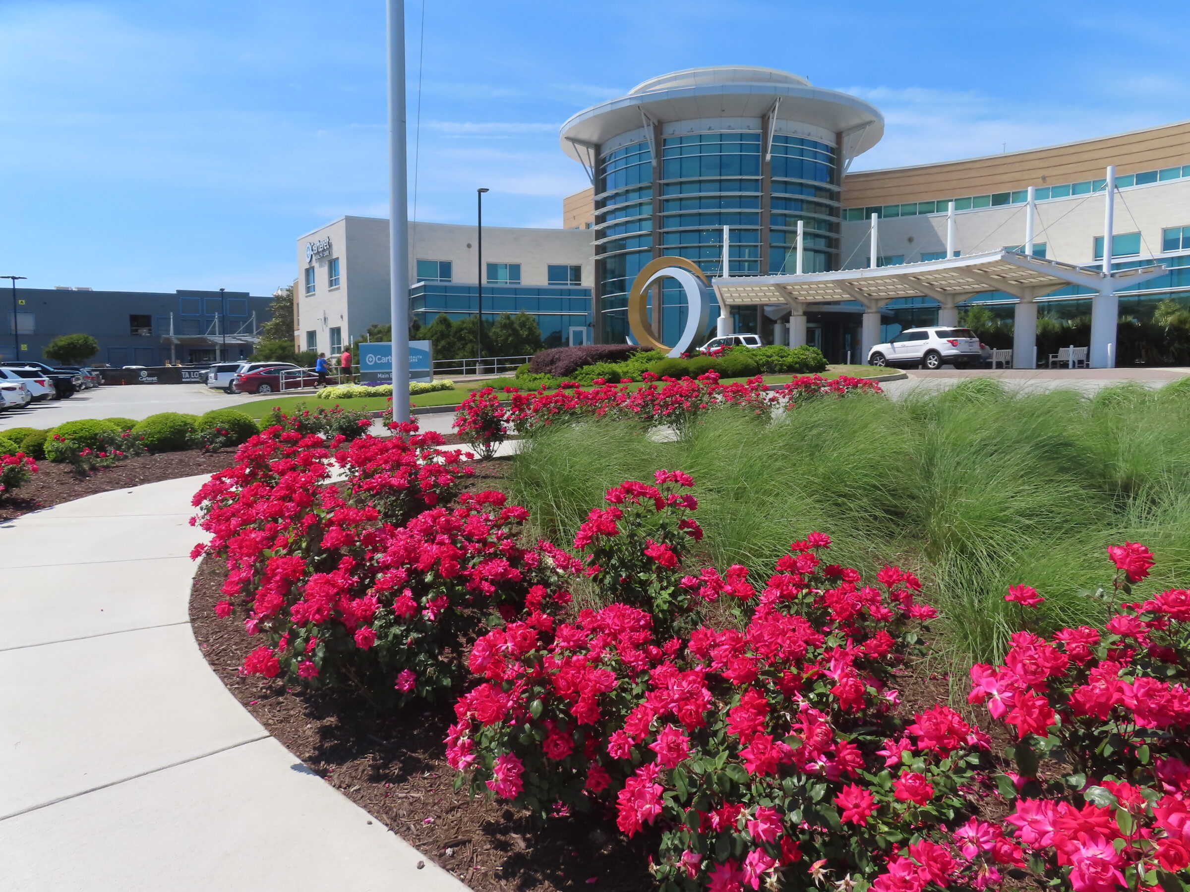 Modern building with a circular entrance, surrounded by vibrant red flowers and green shrubs under a clear blue sky, featuring nearby parked cars.