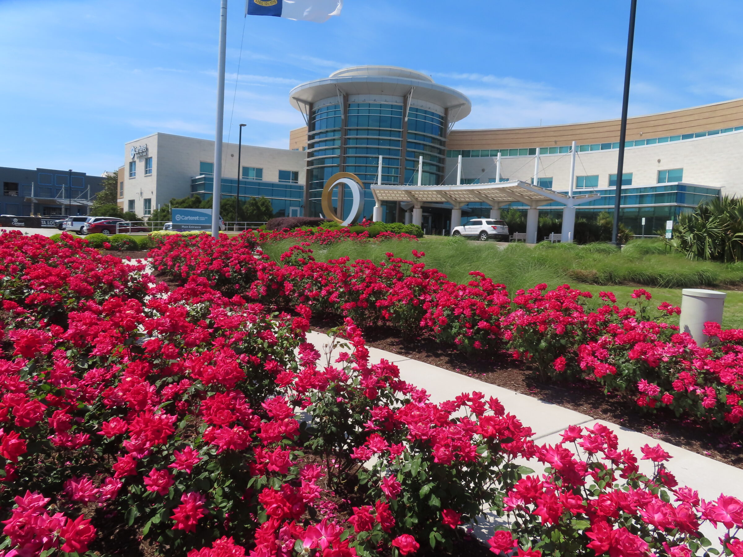 Modern building with curved glass facade, surrounded by vibrant red roses under a clear blue sky. A flagpole stands prominently near the entrance.
