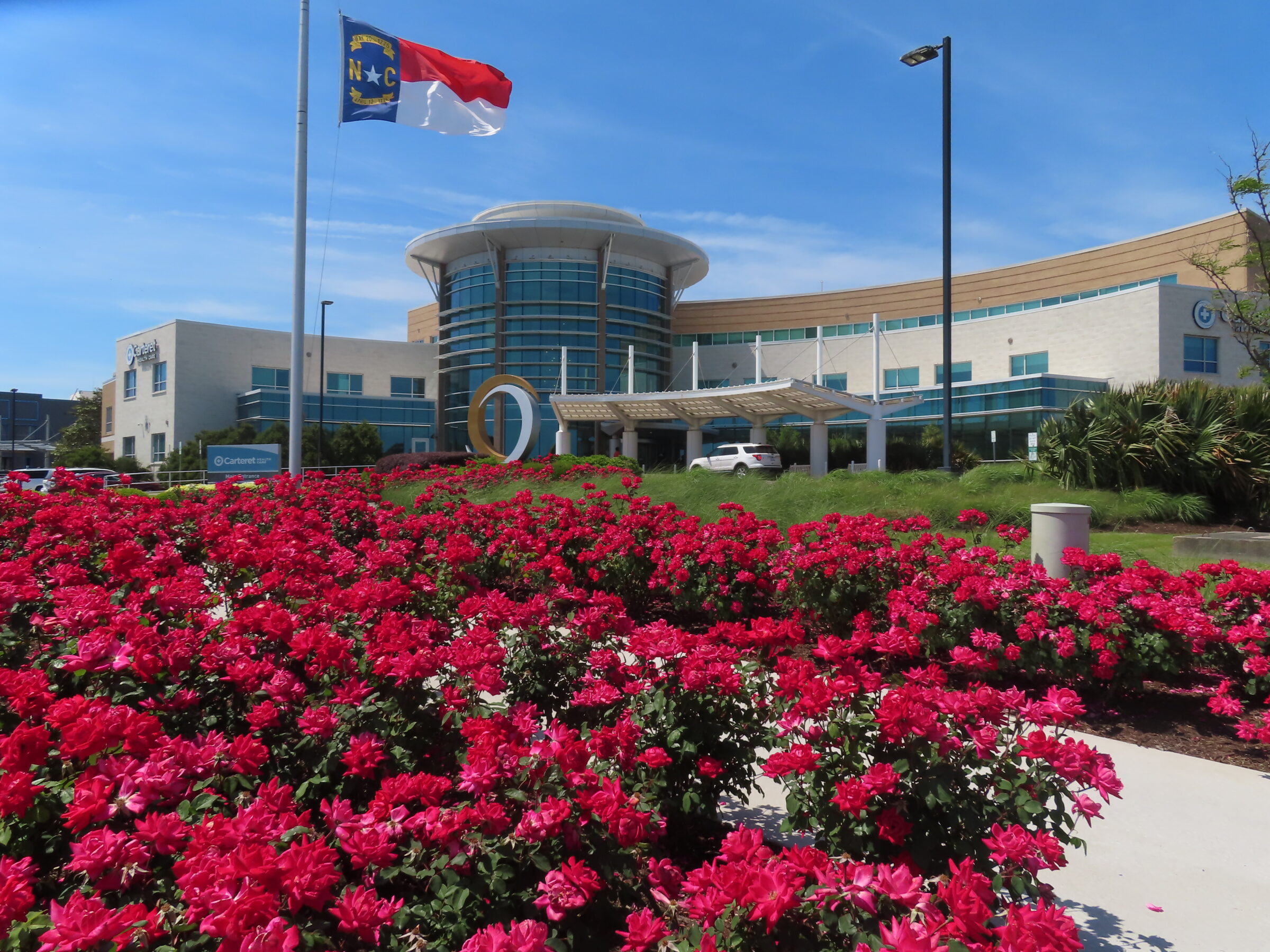 A modern building with glass facade behind vibrant red flowers. North Carolina flag waves prominently in the foreground under a clear sky.