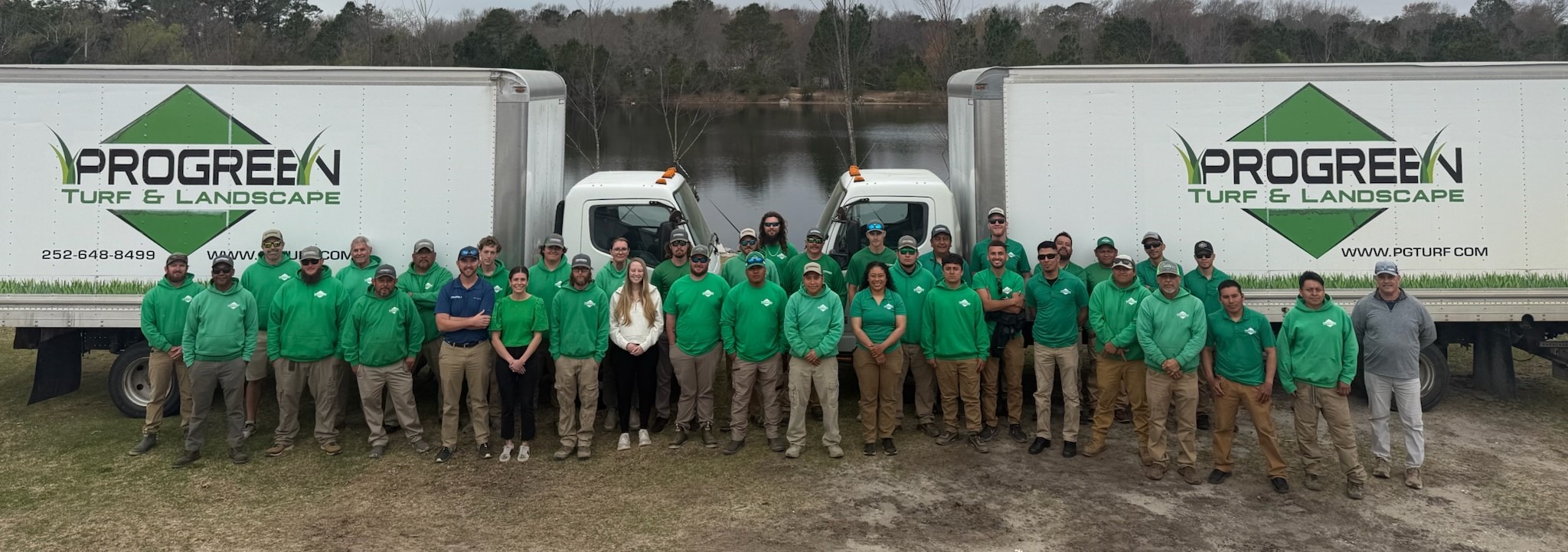 Group of people in green shirts stand between two "Progreen Turf & Landscape" trucks, with a lake and trees in the background.