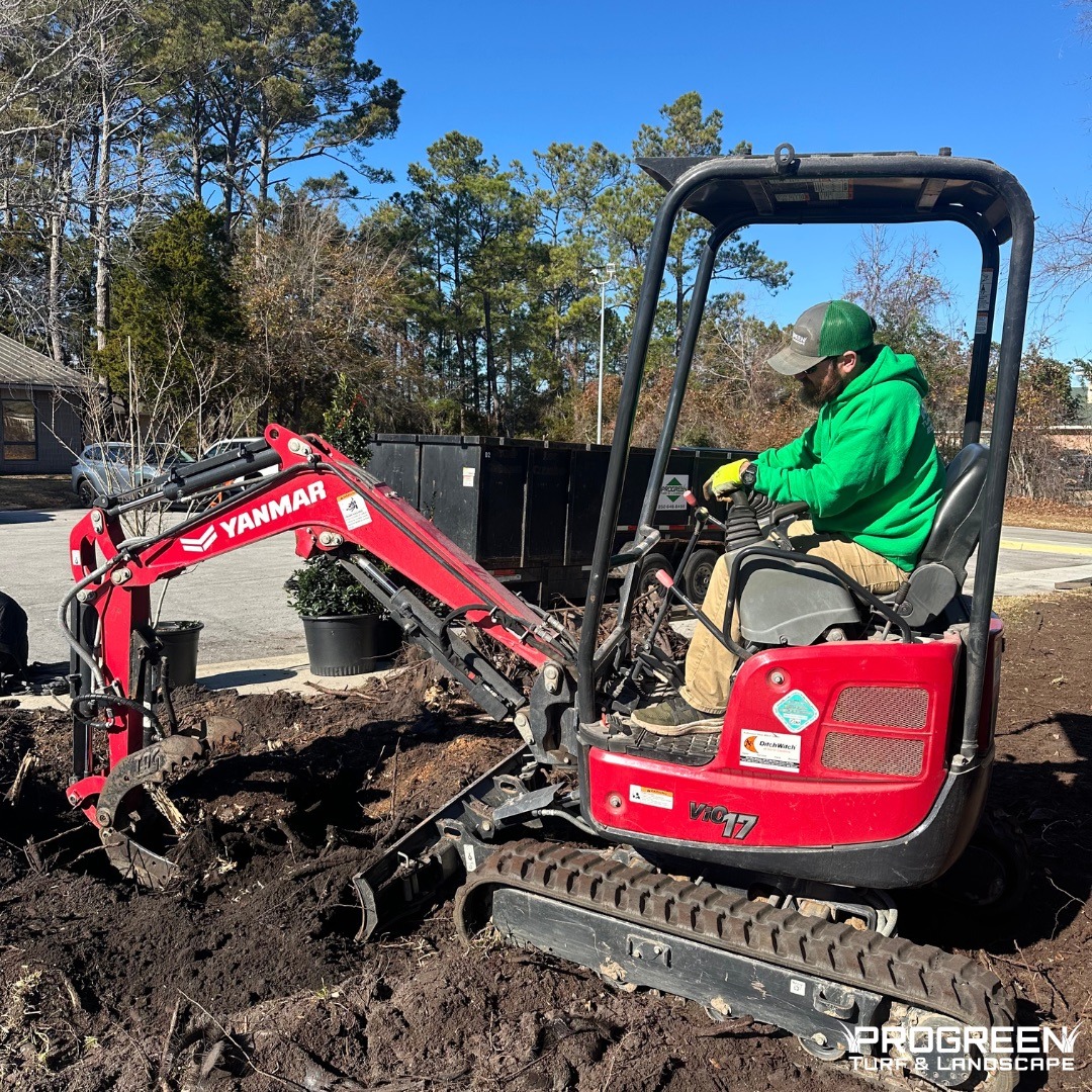A person operates a red mini excavator to dig soil in a landscaped area, surrounded by trees and potted plants under a clear blue sky.