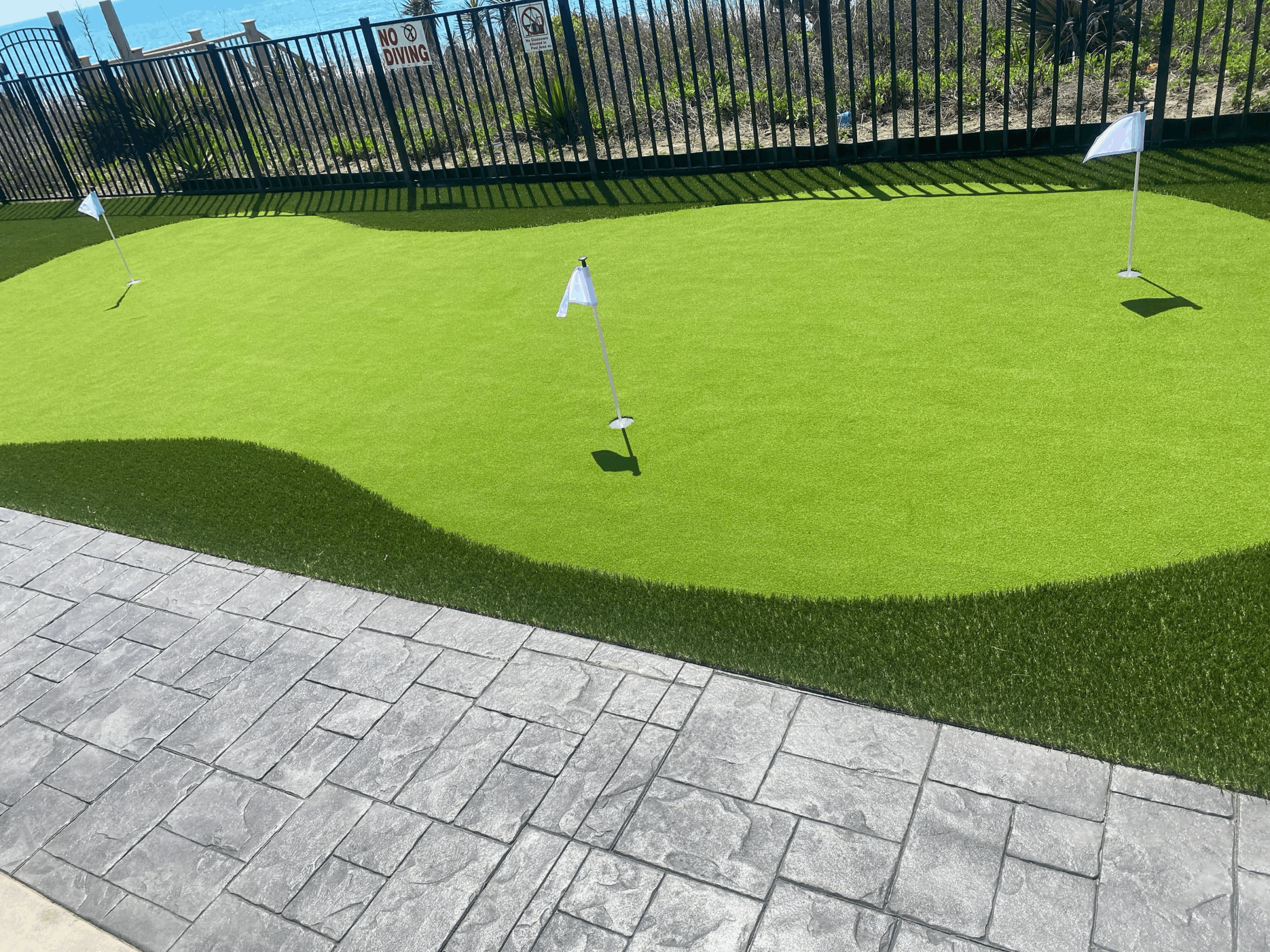 A small golf putting green with three flags is beside a textured stone path and metal fence under a clear blue sky.