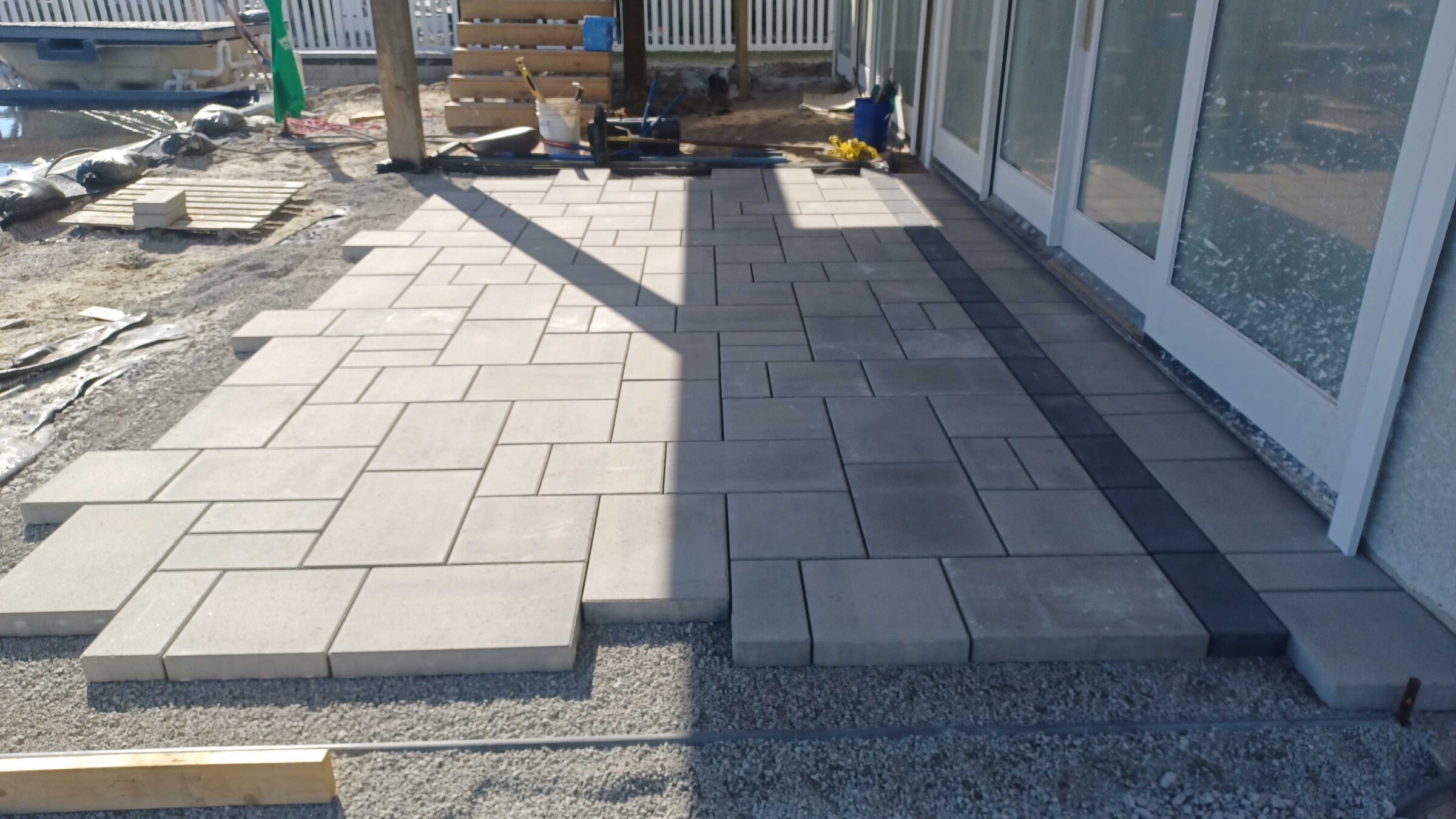 A newly laid patio with geometric paver design beside a building, surrounded by construction materials and a partially fenced yard.