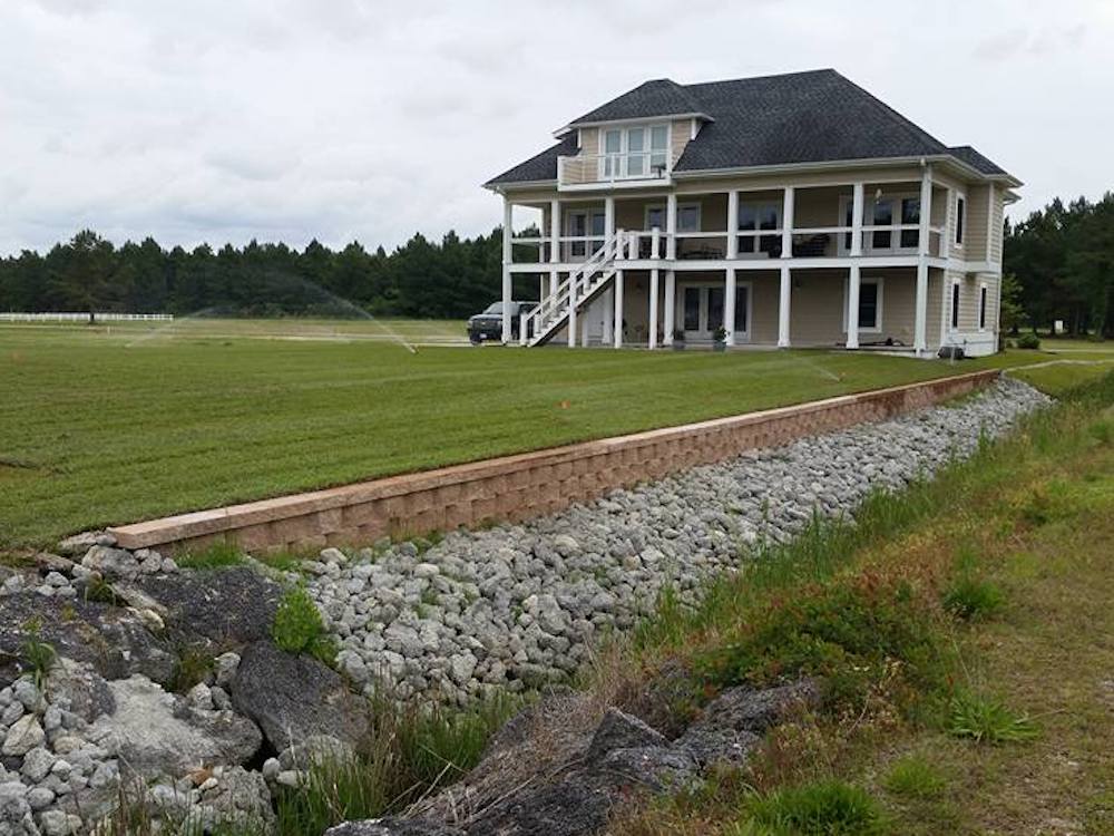 Two-story house with a spacious lawn, rock drainage, and surrounding trees. Sprinkler system waters the grass under a cloudy sky. Peaceful rural setting.