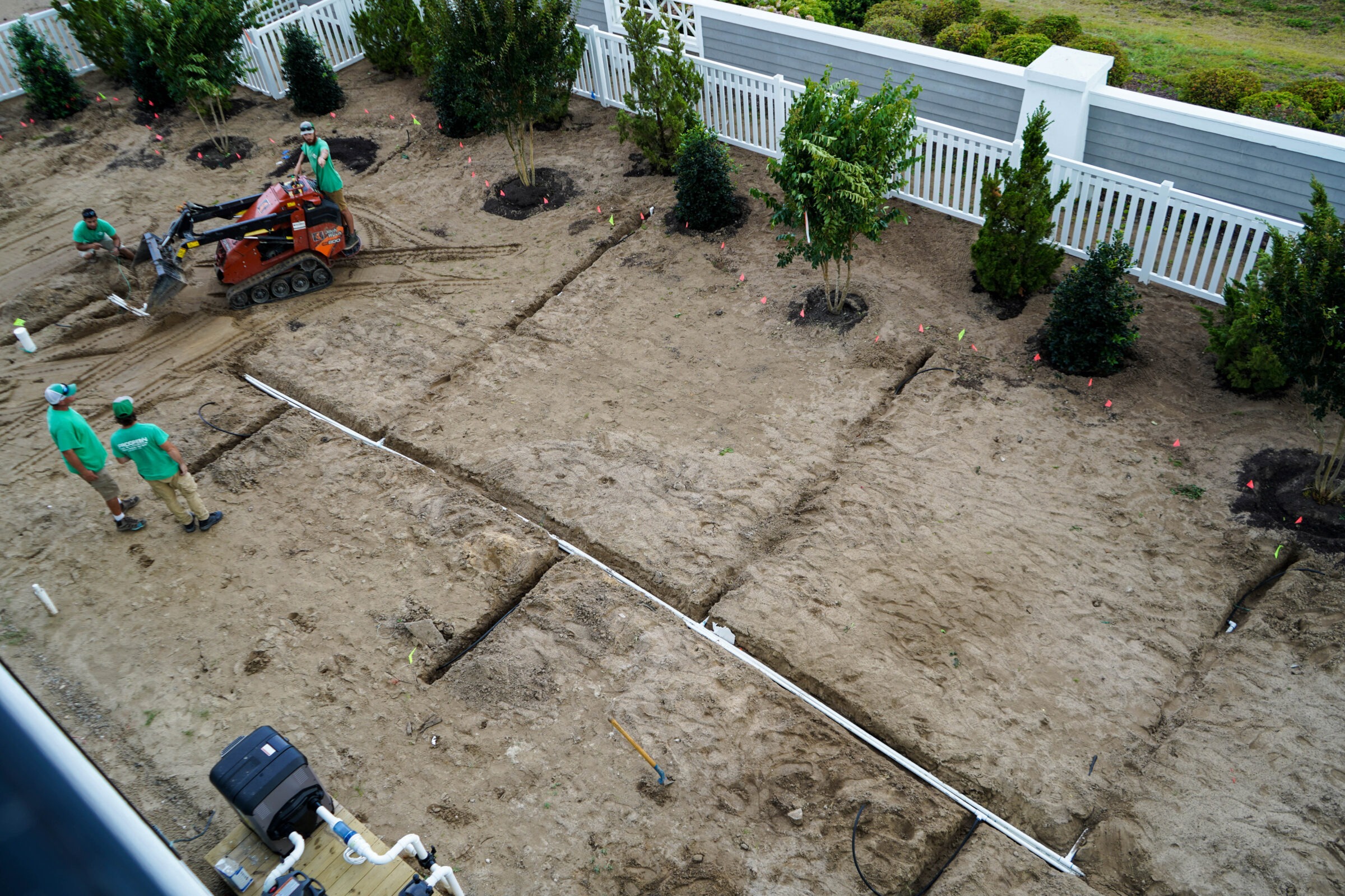 A group of people in green shirts work on landscaping, using machinery to dig trenches near a white fence and newly planted trees.