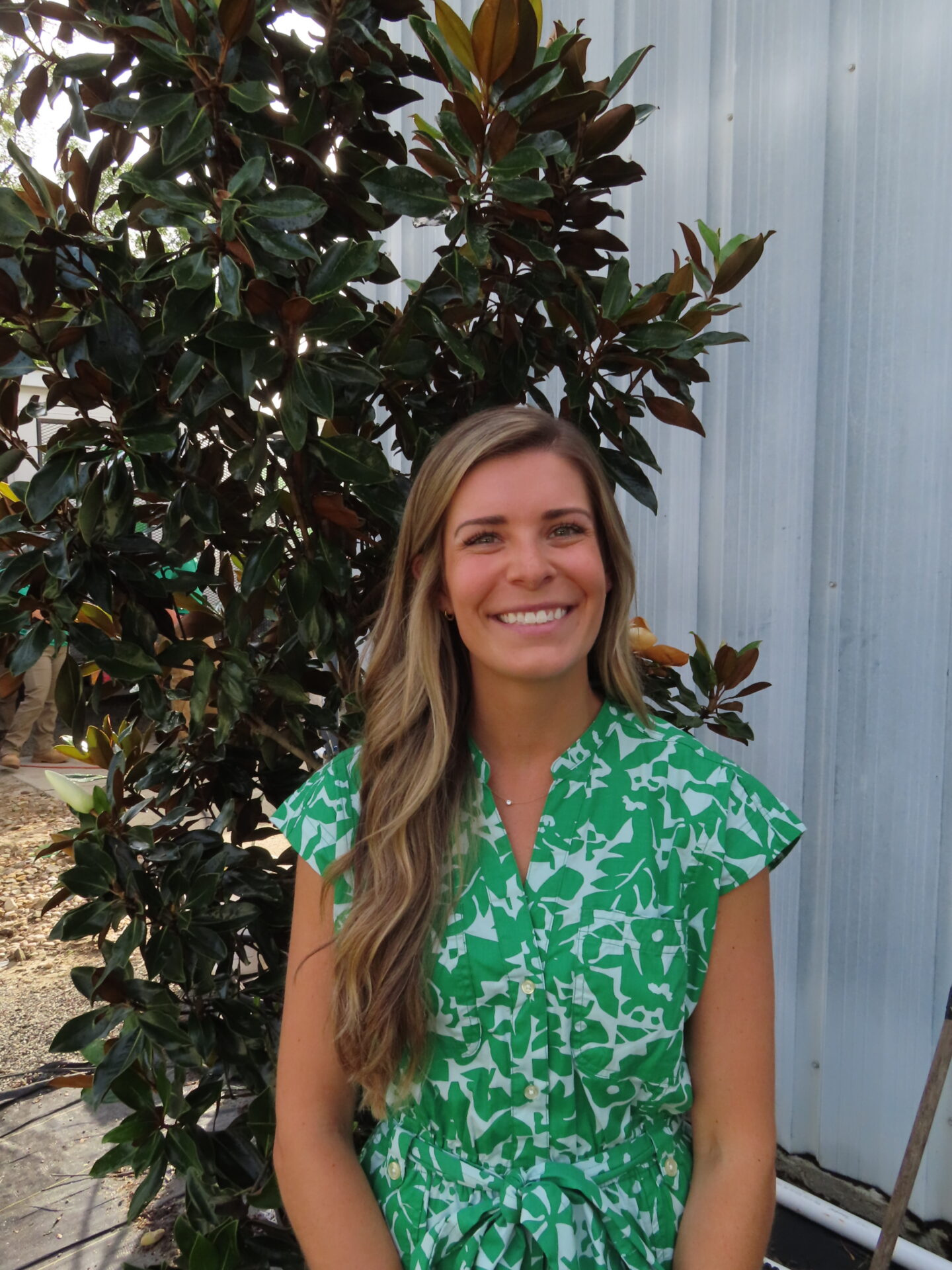 A person in a green patterned dress smiles in front of lush foliage and a corrugated metal wall, creating a vibrant outdoor scene.