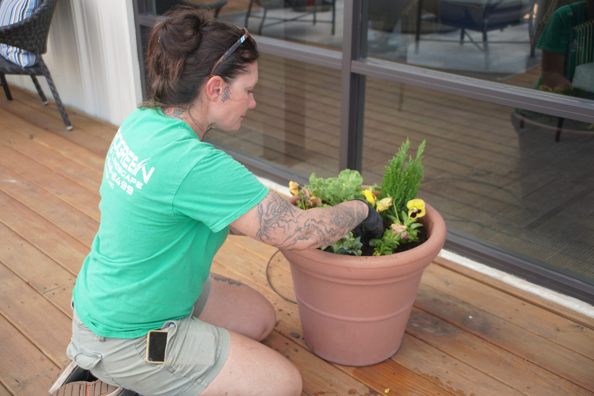 A person with tattoos and green shirt tends to a large planter on a wooden deck. Reflection in windows shows another person.