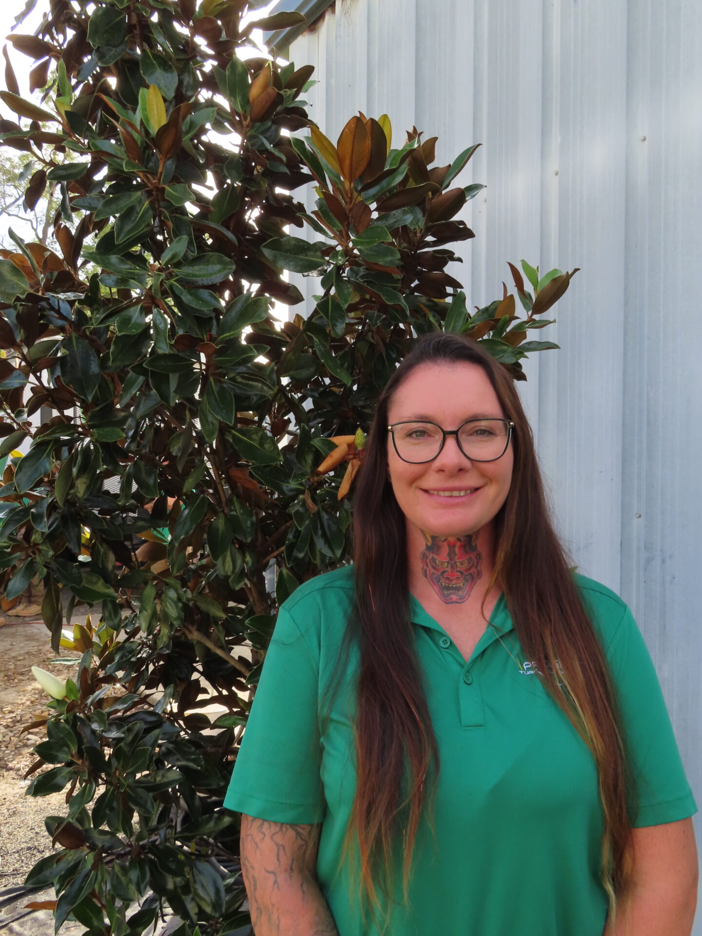 A person in a green shirt stands beside a tree, smiling, with a gray building in the background on a sunny day.