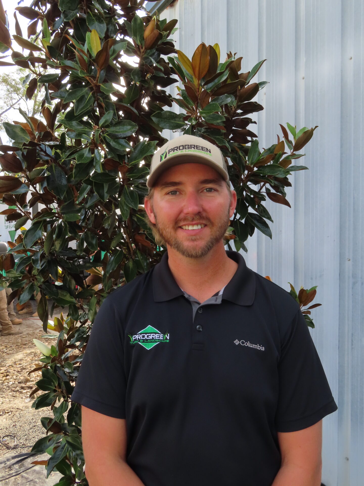 A person wearing a "PROGREEN" cap and black polo stands in front of a magnolia tree and metal wall, outdoors.