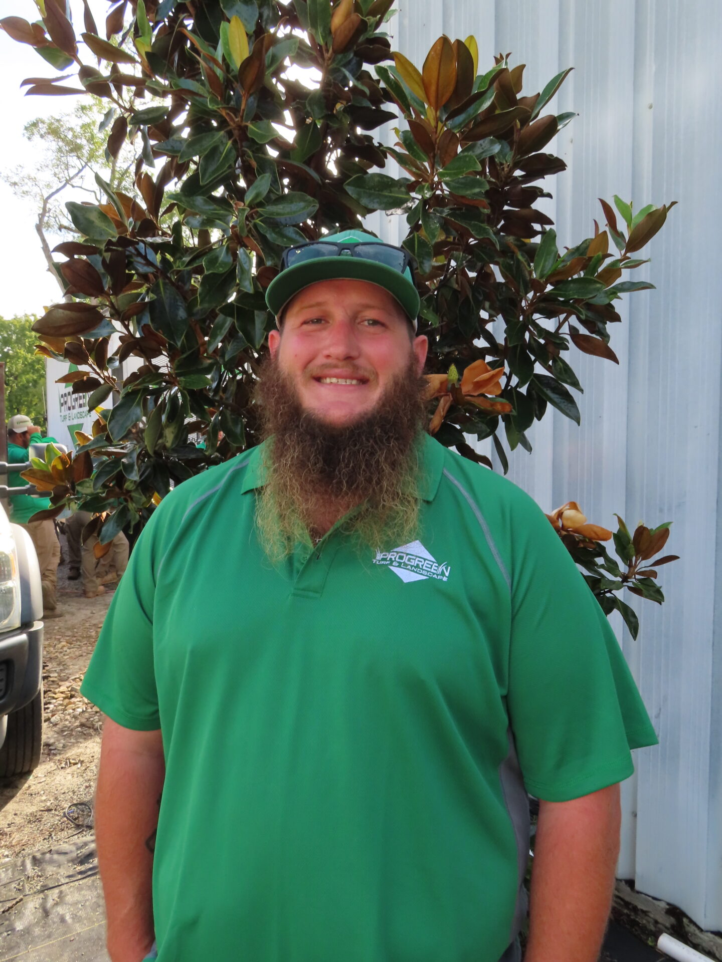 A person with a beard wearing a green shirt and cap smiles by a tree, against a corrugated metal wall. No recognizable landmarks.