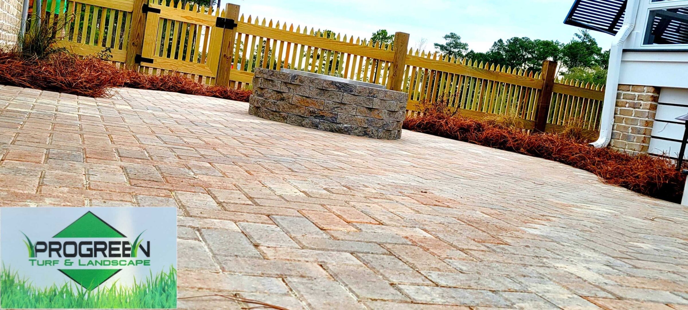 A neatly designed brick patio with a circular stone fire pit bordered by mulch and wooden fence, featuring "ProGreen Turf & Landscape" logo.