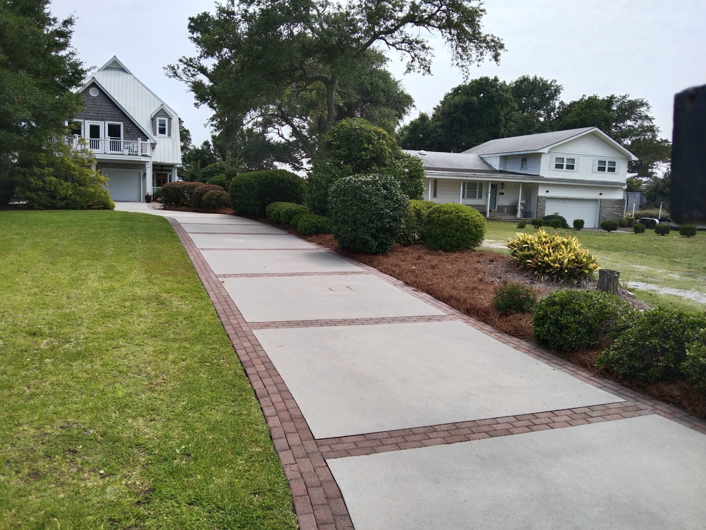 A residential area with two modern houses, manicured lawns, and a paved driveway, surrounded by lush greenery and mature trees.