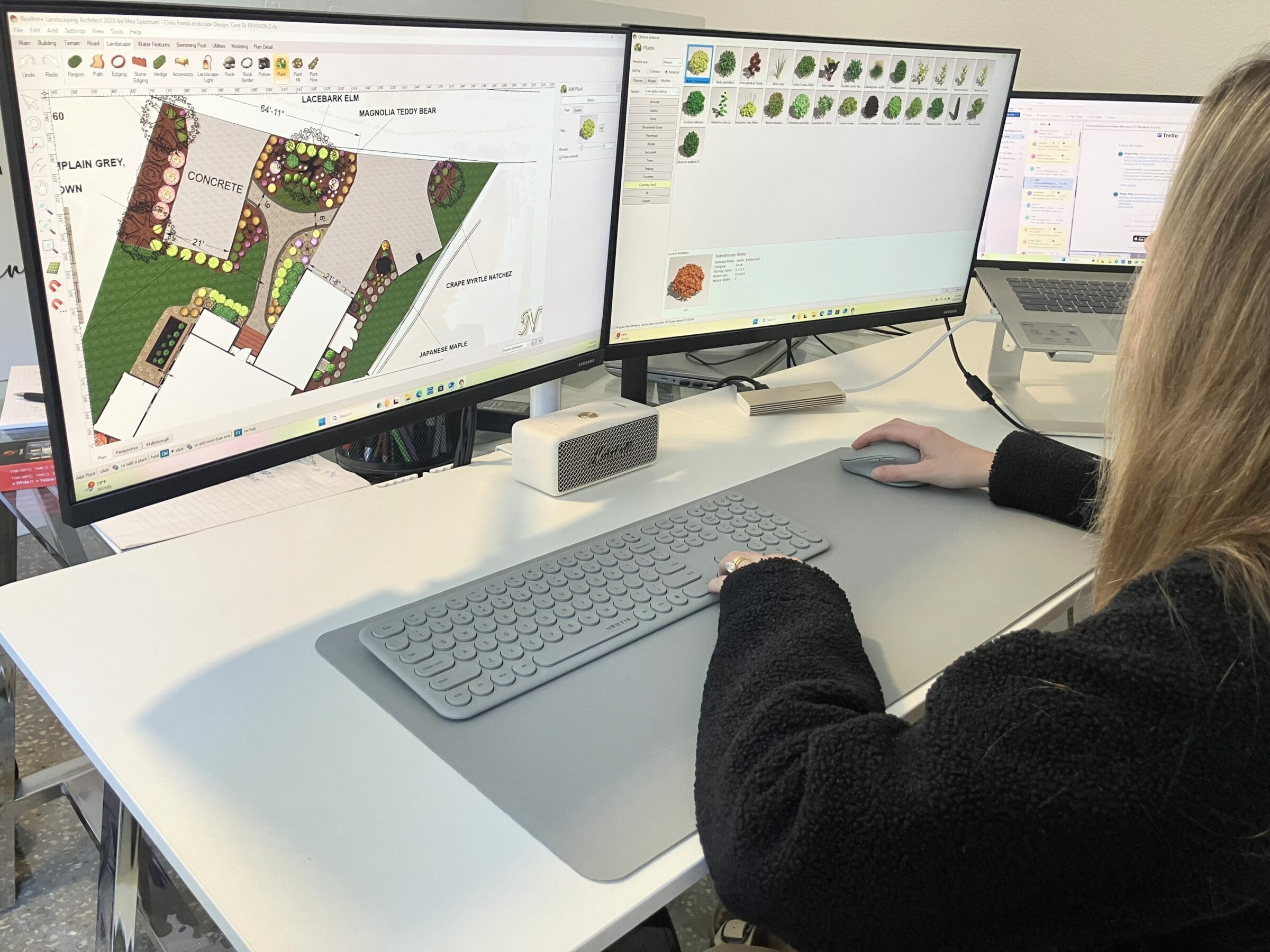 A person uses a computer with dual monitors to design a landscape plan, surrounded by modern office equipment on a sleek desk.