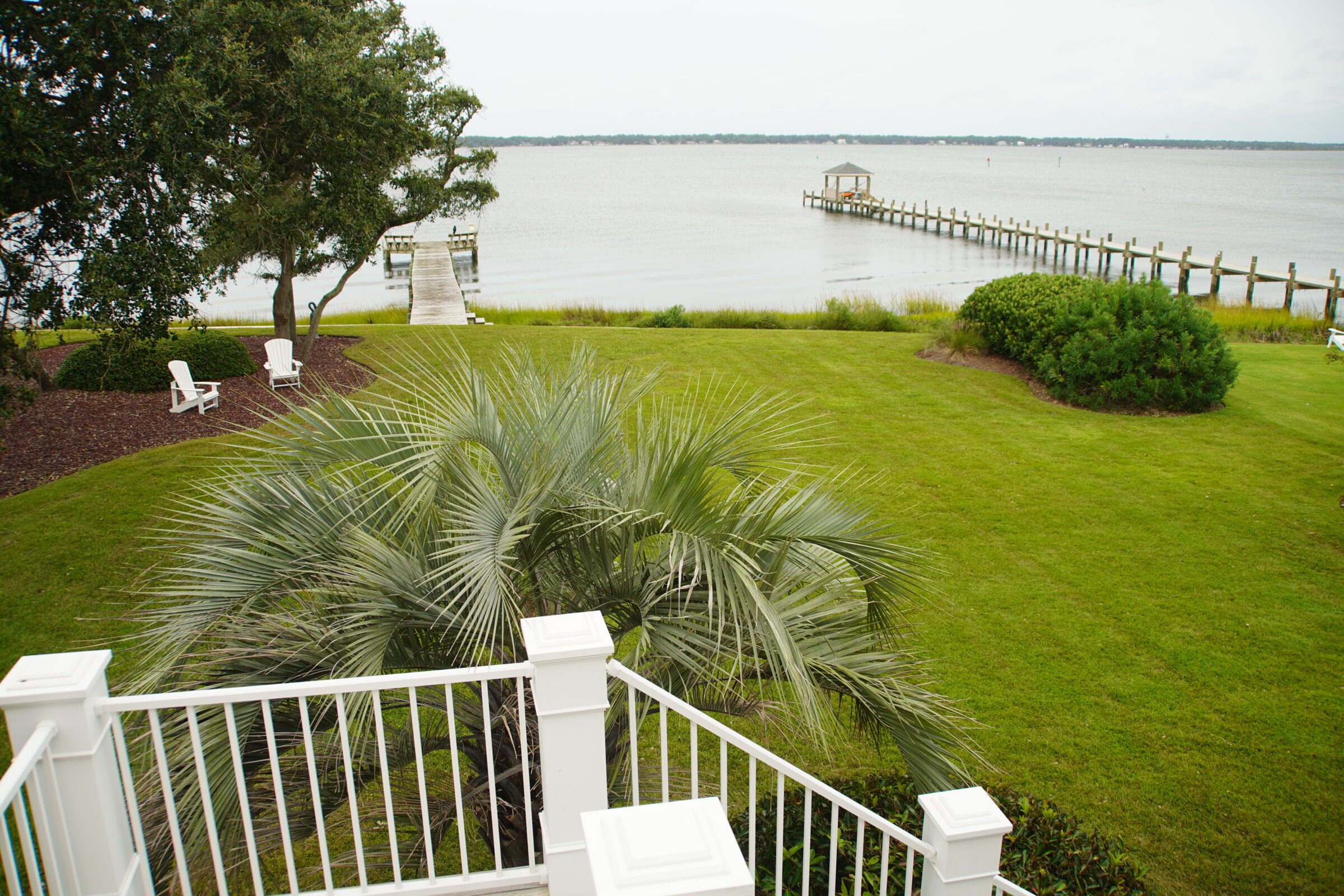 Scenic waterfront view with green lawn, palm tree, two white chairs, and a long wooden pier extending into the calm water.
