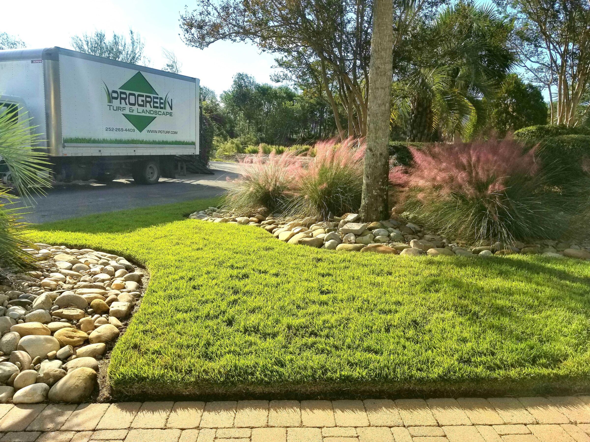 A neatly manicured garden with vibrant grass, decorative rocks, and ornamental pink grasses. A Progreen Turf & Landscape truck is visible.