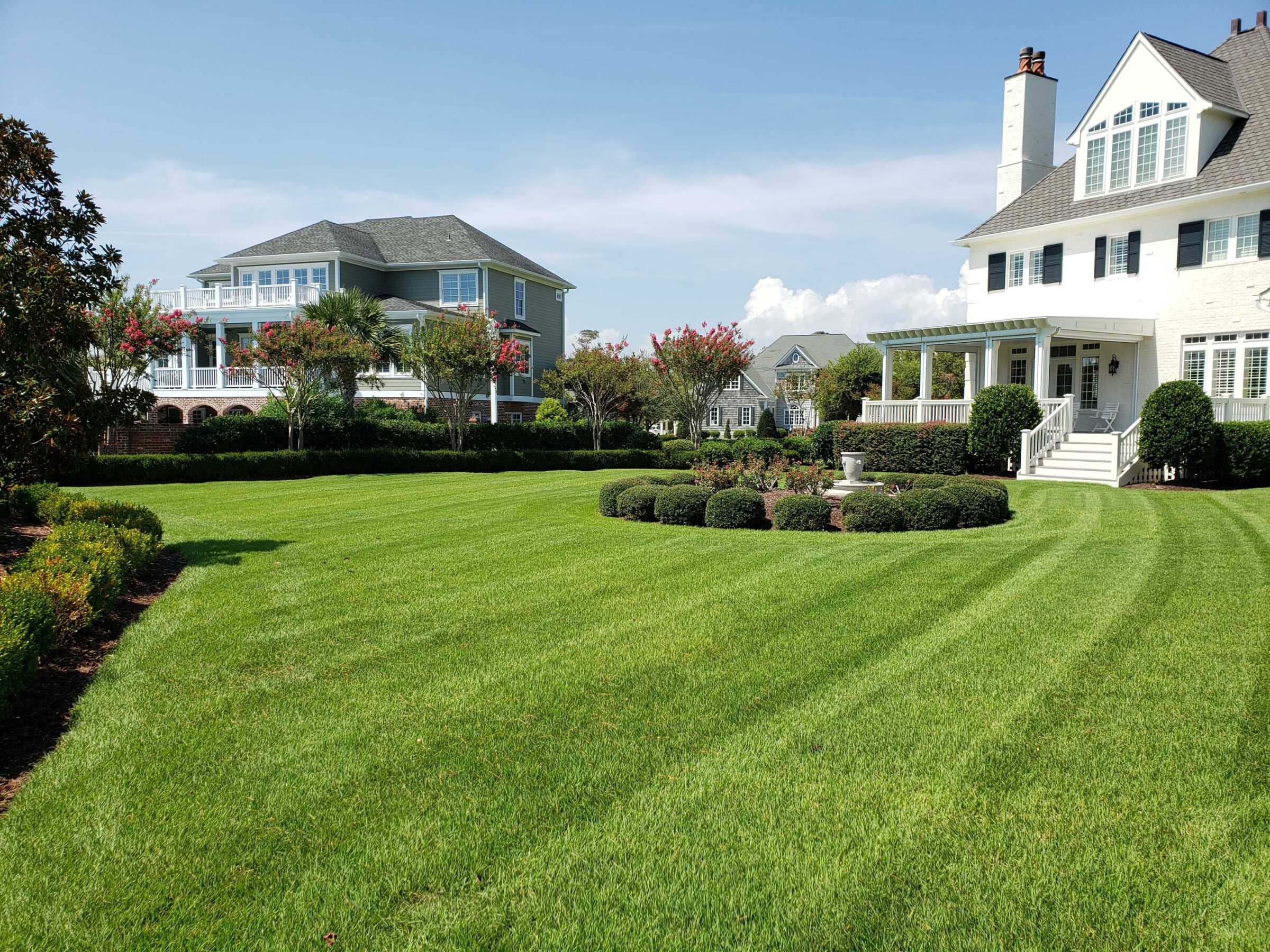 Neatly manicured lawn with vibrant green grass, a white house, trimmed hedges, and a small fountain in a peaceful neighborhood setting.
