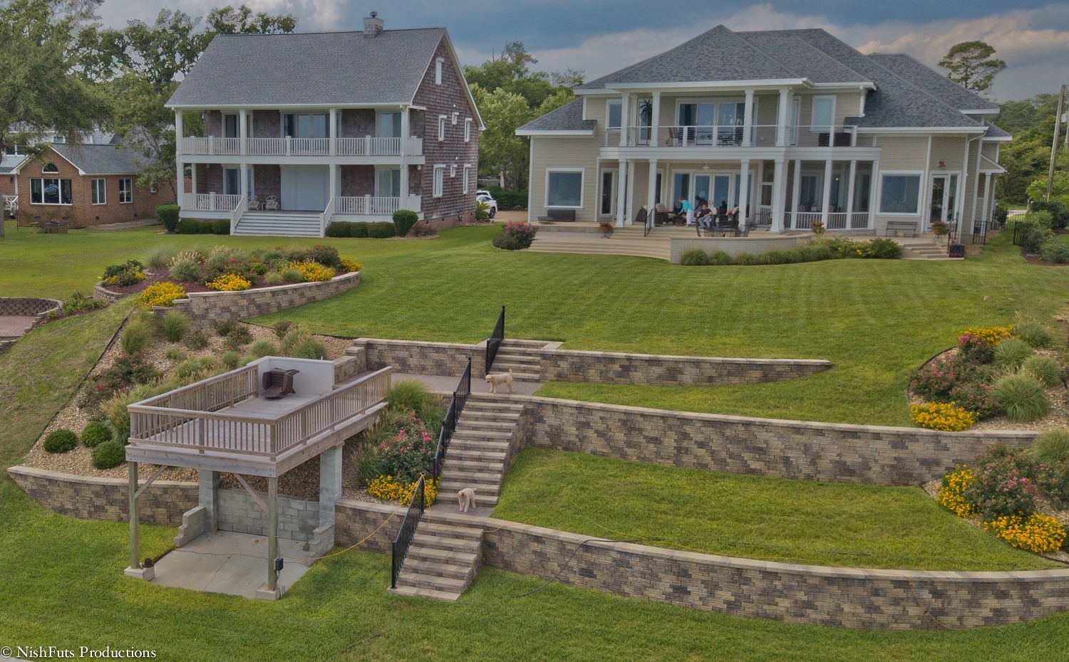 Two large houses with manicured lawns. People sit on the porch, while dogs walk down terraced landscaping with flowers and a small deck.