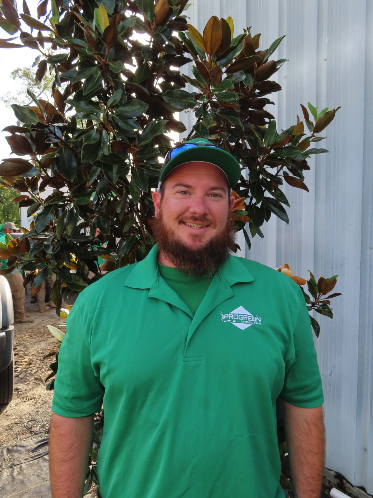A person in a green shirt and cap stands in front of leafy plants, beside a metal wall. No recognizable landmarks or historical buildings.