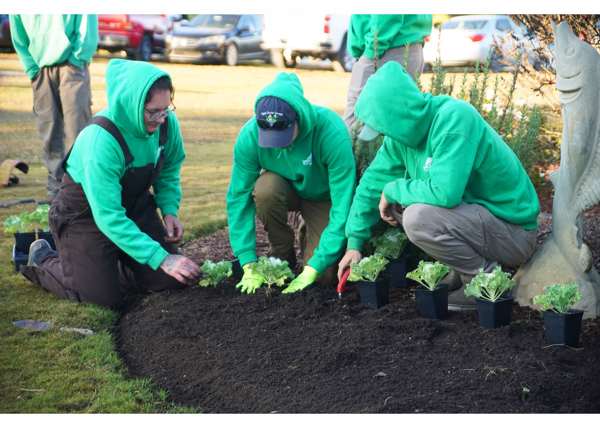 Three people wearing green hoodies plant seedlings in dark soil, with a fish statue nearby. Cars are parked in the background.