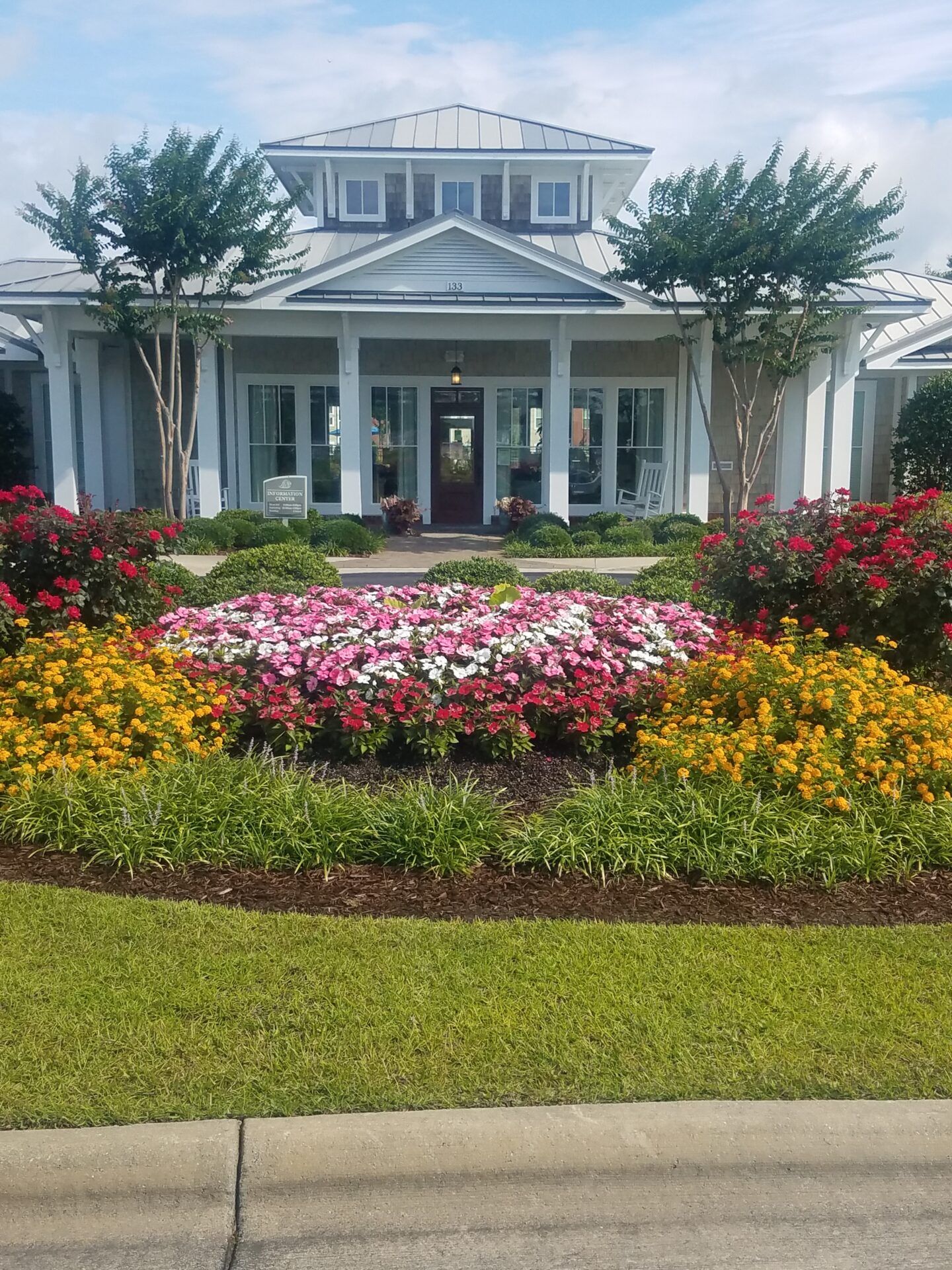 A charming house with a white facade, surrounded by vibrant flower gardens and manicured shrubs, under a blue sky with scattered clouds.