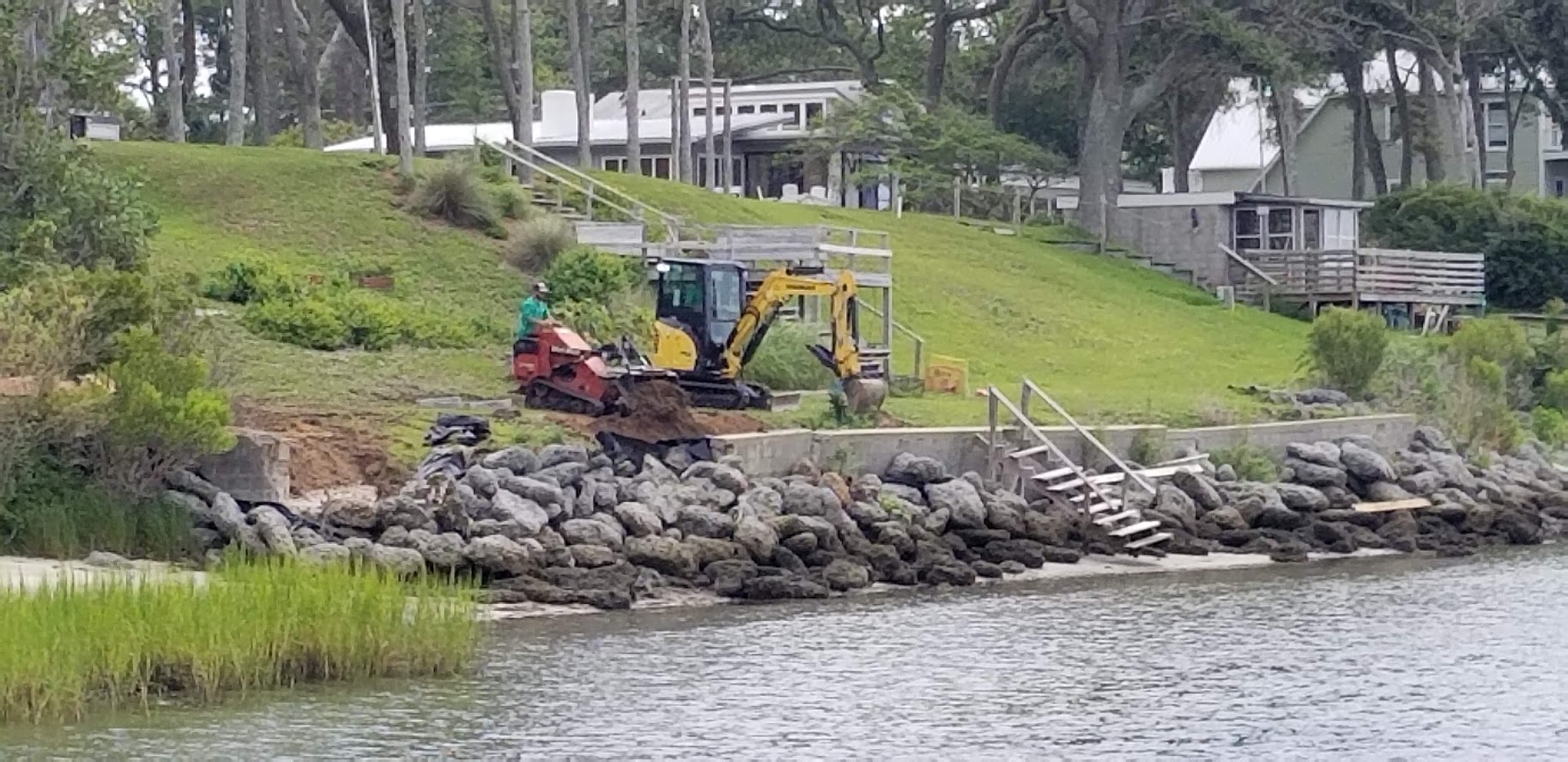 A person operates machinery beside a rocky shoreline, with a grassy slope and modern buildings in the background, surrounded by trees.