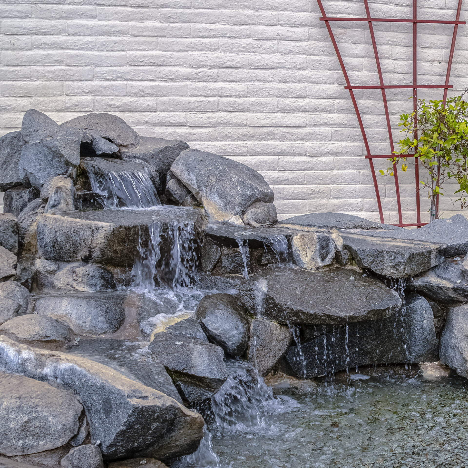 A small stone waterfall with cascading water against a brick wall, adorned with a trellis and a green plant.