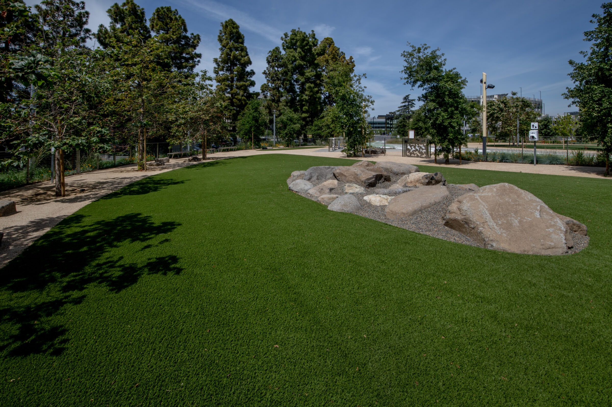 A serene park scene with lush grass, large rocks, and tall trees under a clear blue sky, featuring walkways and distant contemporary buildings.