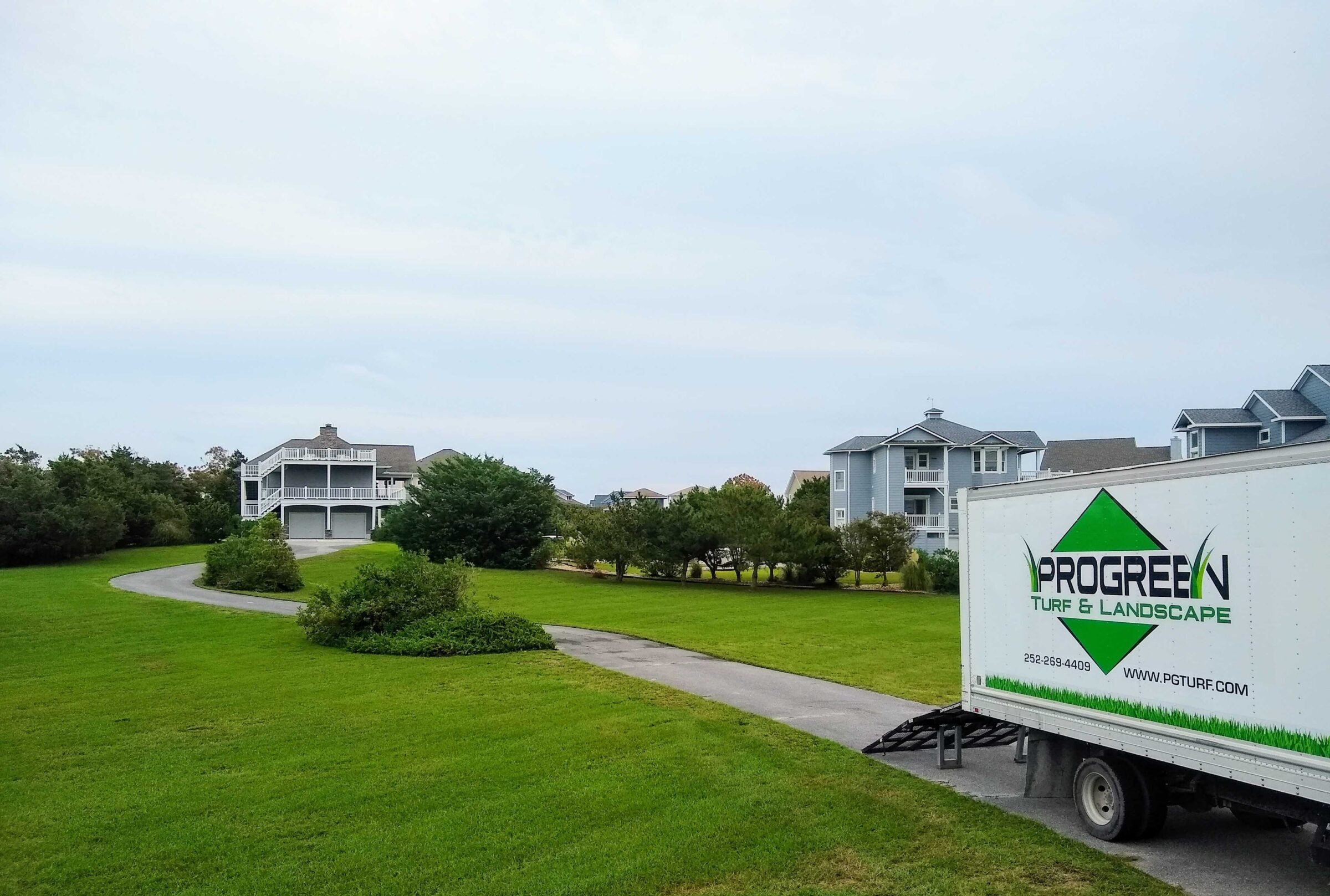 A suburban landscape features a neatly mowed lawn, residential houses, and a ProGreen Turf & Landscape truck on a curving driveway.