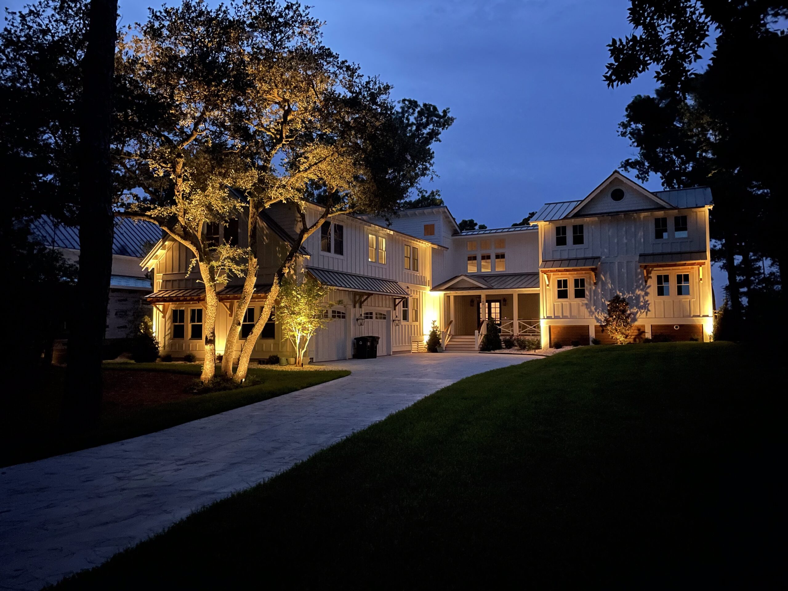 A large, well-lit house with a long driveway is surrounded by trees in a peaceful, twilight setting.