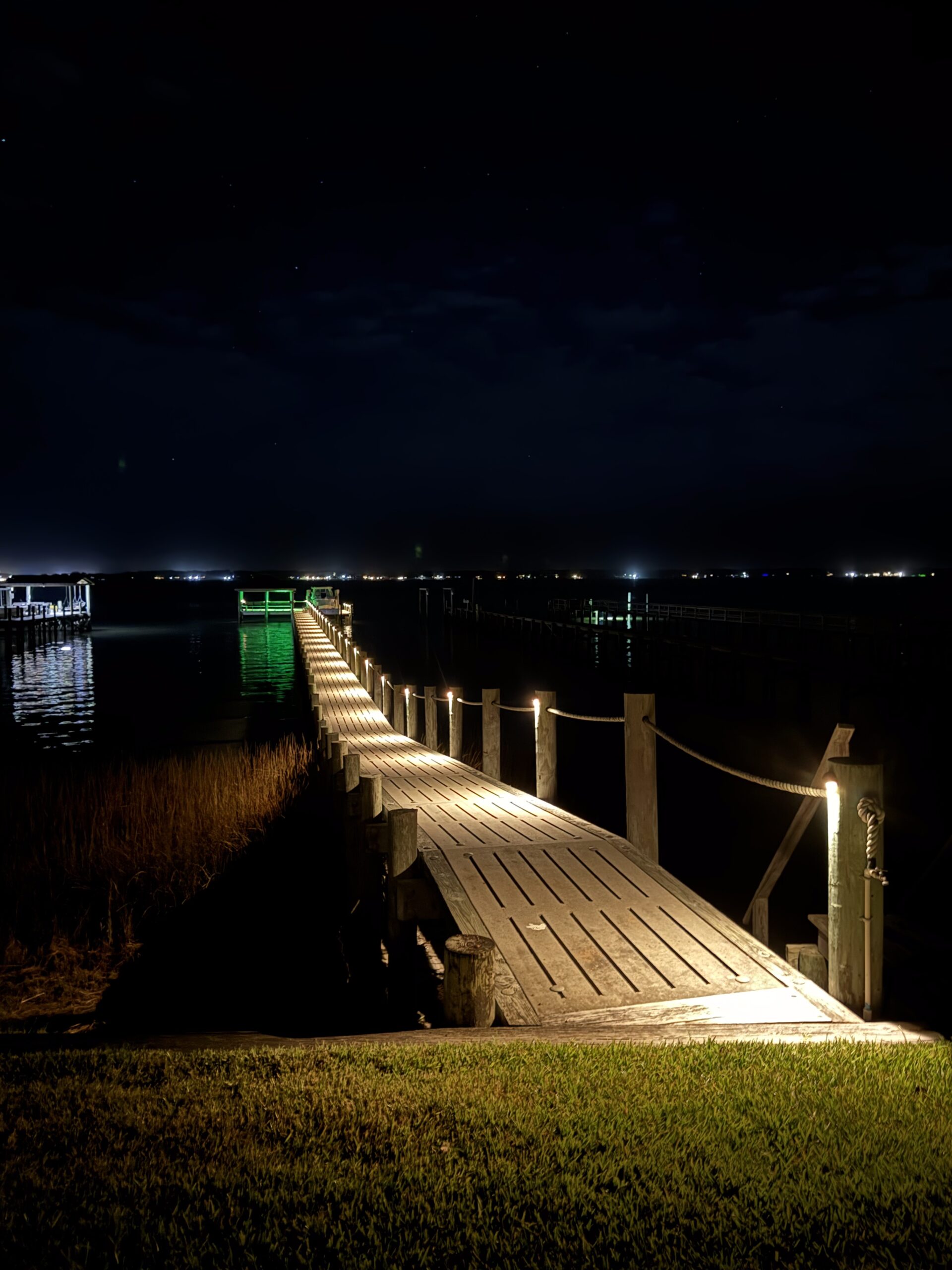 A wooden pier with warm lights extends into a dark, starry sky over a calm, reflective water body at night.