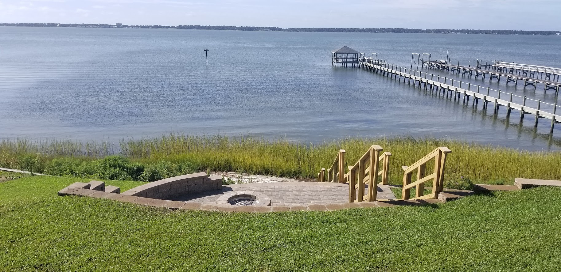 A tranquil waterfront scene with a dock, grassy slope, and circular stone fire pit, overlooking a calm, wide body of water.